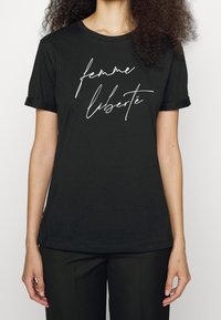 Camiseta de algodón negra con mangas enrolladas, que presenta texto en cursiva blanca "femme liberté" en la parte delantera. Diseño simple de cuello redondo.