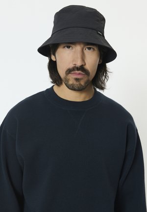 Schwarzer Bucket-Hut mit glatter Textur und leichtem Glanz. Kombiniert mit einem dunkelblauen Sweatshirt mit Rundhalsausschnitt und normaler Passform.