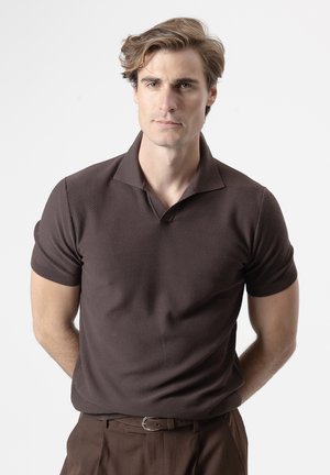 Joven con cabello castaño claro, vistiendo una camiseta tipo polo de manga corta marrón oscuro y pantalones a juego, de pie con las manos detrás de la espalda.