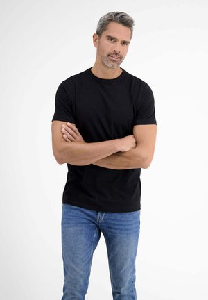 Man with crossed arms wearing a black t-shirt and blue jeans, standing against a plain white background.