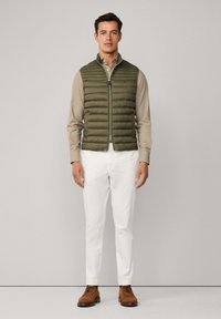 Olivi vihreä pehmustettu liivi vetoketjulla, päällä vaaleanruskea paita, valkoiset housut ja ruskeat kengät, neutraalissa taustassa.