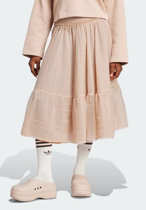 Osoba nosící béžovou midi sukni s elastickým pasem, bílé ponožky Adidas a béžové platformové nazouvací boty, stojící na světlém pozadí.