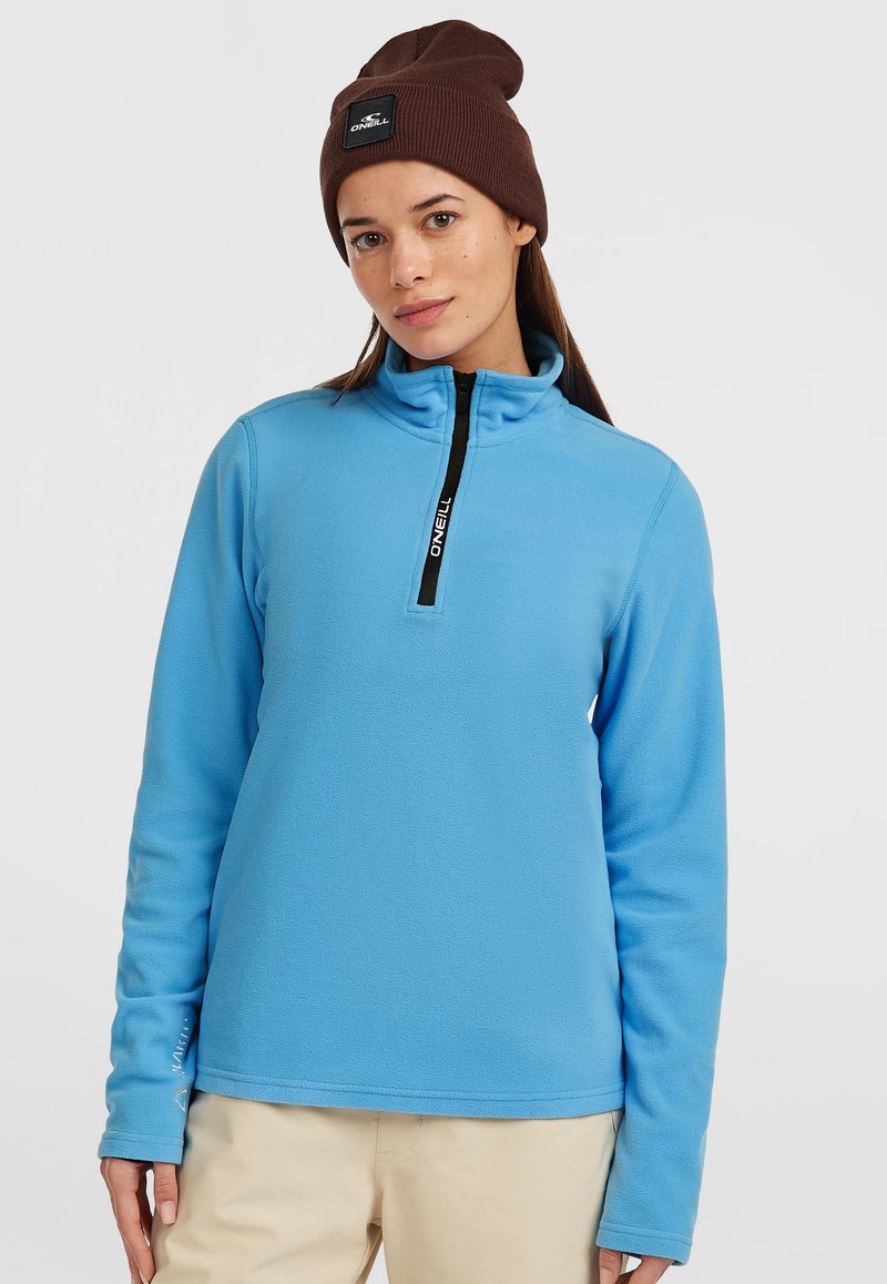 Lyseblå fleece pullover med en kvart lynlås, sort lynlåsdetalje og lange ærmer, båret med en brun strikket hue.
