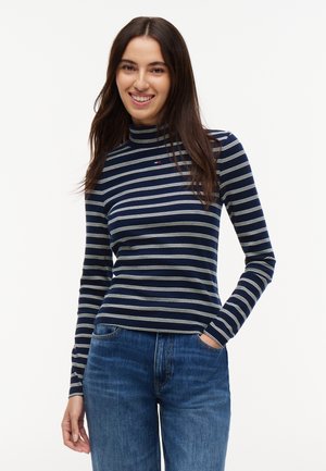 Jeune femme aux longs cheveux foncés sourit, portant un haut à manches longues rayé bleu marine et blanc et un jean bleu, main dans la poche.