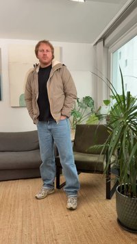 helepruun lukuga jakk valge voodriga, must särk all, sinised teksad ja hallid tossud. Siseala taimedega ja diivaniga.