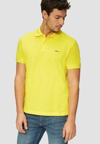 Gelbes Poloshirt aus Baumwolle mit klassischem Kragen, kurzen Ärmeln und einem kleinen schwarzen Logo auf der Brust, kombiniert mit blauen Jeans.