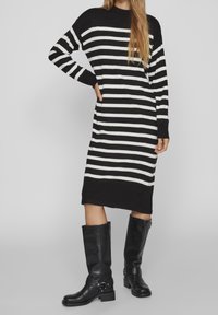 Randig, långärmad klänning i svart och vitt med en avslappnad passform. Bärs med knähöga svarta läderstövlar med dekorativ hårdvara.