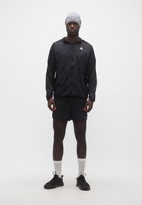 Άνδρας που στέκεται, ντυμένος με μαύρο αθλητικό μπουφάν, μαύρο σορτς, γκρι σκούφο, λευκές κάλτσες και μαύρα παπούτσια τρεξίματος, μπροστά σε ουδέτερο φόντο.