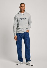 Grijze hoodie met marineblauwe geborduurde tekst "Pepe Jeans," gecombineerd met losse blauwe denim broek en grijze sportschoenen. Glad stoftextuur.