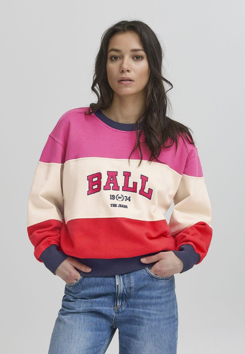 Flerfarget hettegenser med rosa, kremfarge og røde seksjoner. Har fet "BALL 1974" tekst, ribbestrikkede mansjetter og en marineblå krage. Avslappet passform.