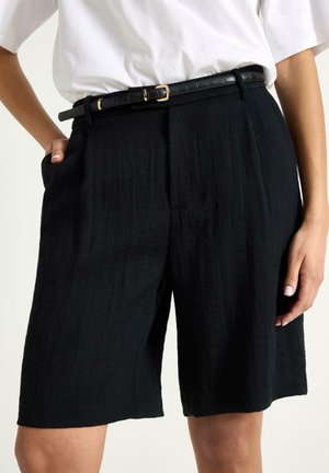 Henkilö pukeutuneena korkeavyötäröisiin mustiin räätälöityihin shortseihin, joissa sivutaskut ja kapea musta vyö, yhdistettynä valkoiseen lyhythihaiseen toppiin.