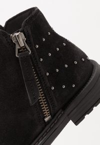 Svart mocka ankelsko med silvernitar, sidodragkedja och strukturerad sula. Funktioner inkluderar en låg klack och förstärkt sömnad längs kanterna.