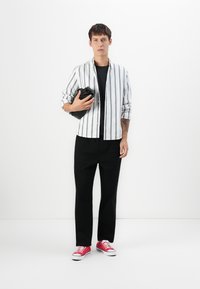 Joven de pie, vistiendo una camisa blanca a rayas sobre una camiseta negra, pantalones negros, zapatillas rojas, sosteniendo una chaqueta negra plegada.