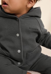 Gray waffle-knit hoodie with a zipper, featuring silver snap buttons and a front pocket, branded with "ALLSAINTS" on the chest.
