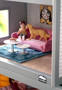 Dollhouse scene featuring a pink velvet sofa, two metallic tables, a blue fur rug, a cartoon art piece, and a child with a golden dog.