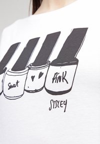 Bílý tričko s grafikou lahviček laků na nehty; barvy zahrnují černou, růžovou a bílou; text obsahuje "Swat", srdce a "PINK", od Sisley.