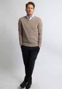 Beige gebreide trui over een lichtblauw overhemd met kraag, gecombineerd met zwarte broeken en donkere schoenen. Staande pose met handen in de zakken.