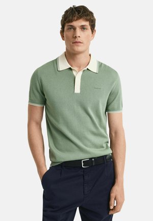 Grön kortärmad polo med krämfärgad krage och randiga detaljer; tyget ser slätt och åtsittande ut, med en subtil logotyp på bröstet.