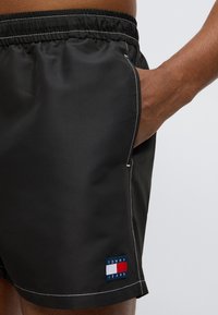 Μαύρα μαγιό σορτς με ελαστική μέση, λευκά ραφές και πλαϊνή τσέπη. Διαθέτουν μια μπλε ετικέτα με κόκκινες και λευκές λεπτομέρειες που αναγράφει "TOMMY JEANS."