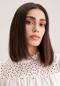 Blusa bianca con motivo ricamato e colletto arricciato. Il modello ha capelli scuri e lisci e trucco neutro, mettendo in risalto lineamenti facciali simmetrici.
