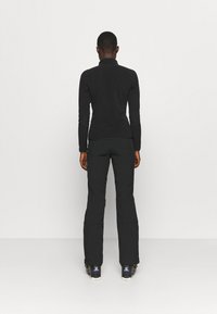 Schwarzes Langarm-Oberteil mit hohem Kragen, kombiniert mit schwarzen Hosen. Das Outfit zeichnet sich durch ein tailliertes Design und eine glatte Textur aus. Schwarze Schuhe vervollständigen den Look.