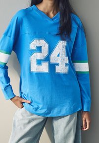Sininen pitkähiha-paita, jossa valkoinen kulunut "24" etupuolella. Hihansuissa vihreä- ja valkoraidalliset yksityiskohdat. Pehmeää puuvillamateriaalia.
