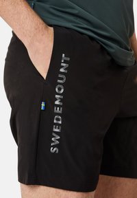 Svarta simshorts med reflekterande "SWEDEMOUNT"-text på sidan och en liten svensk flagga nära fickan. Slät tygtextur.