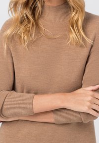 Svetr s vysokým límcem v světle hnědé barvě, s žebrovanými manžetami a límcem, charakterizovaný měkkou texturou a přiléhavým střihem.