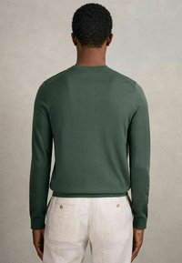 Dunkelgrüner langärmeliger Pullover aus weichem Stoff, mit Rundhalsausschnitt und eng anliegenden Manschetten, kombiniert mit hellen Leinenhosen.