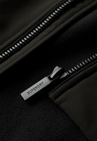 Zwarte ritssluiting met "SUPERDRY" gegraveerd op de trekker, naast een zachte stof oppervlakte, met een contrasterend ritsontwerp.