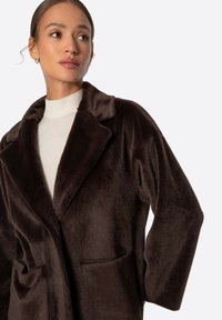 Abrigo de pelo sintético marrón con cuello muesca, dos bolsillos frontales y una textura suave. El diseño presenta un corte relajado y está desabrochado.