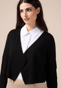 Svart kortärmad cardigan med V-ringning, tillverkad av texturerad tyg, bärs över en vit skjorta med krage. Mjuk och bekväm passform.