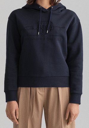 Granatowa bluza z kapturem ze ściągaczami i wytłoczonym logo "GANT American Sportswear", noszona z beżowymi plisowanymi spodniami.