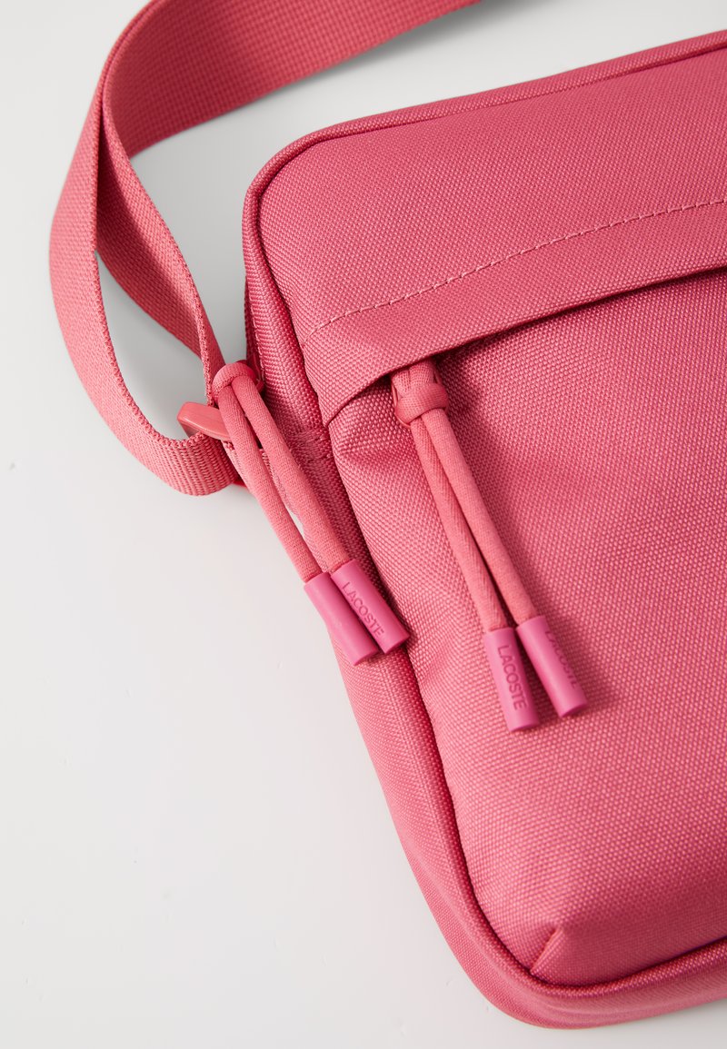 Sac messager rose avec un tissu texturé, fermeture éclair et tirettes assorties arborant le logo de la marque. Sangle ajustable incluse.