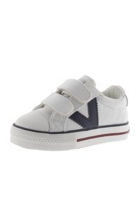 Zapatilla de lona blanca con acento en forma de V en azul marino, doble banda de velcro, punta de goma y franjas rojas y azul marino a lo largo de la suela.