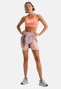 Roxy sportbeha in koraal met wit logo, gecombineerd met gestreepte lavendel short. Een paarse sweat shirt is om de taille geknoopt. Sport schoenen zijn gedragen.