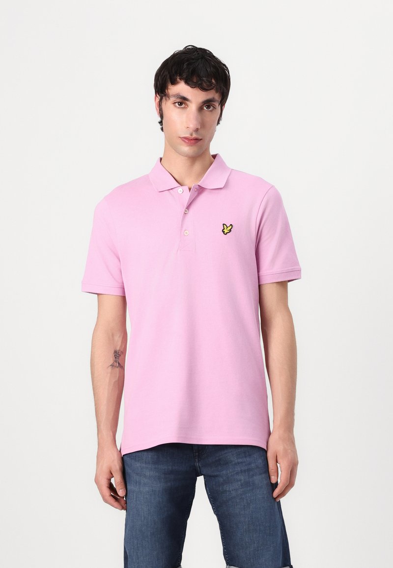 Rosa Poloshirt aus Baumwolle mit einem gelben gestickten Logo auf der linken Brust. Kurze Ärmel, flacher Kragen und gerader Schnitt.