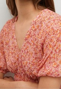 Blouse légère à motif floral avec une base rose, ornée de petites fleurs orange et jaunes, manches ballons, et un décolleté en V avec un détail de taille smockée.
