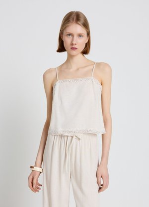 Jeune femme aux cheveux blonds courts portant un haut blanc sans manches avec une bordure en dentelle et un pantalon blanc assorti, debout devant un fond clair.