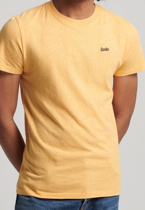 Homme portant un t-shirt jaune à manches courtes avec un petit logo "SuperDry" sur la poitrine, associé à un jean bleu, debout devant un fond uni.
