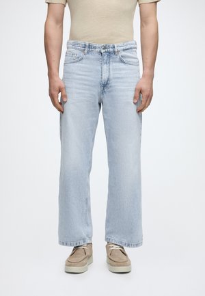 Mężczyzna w jasnoniebieskich jeansach szerokich nogawkach, beżowym topie z krótkim rękawem i beżowych casualowych butach, stojący na jednolitym białym tle.