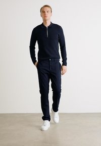 Mornarska polo majica na zatvori, pripijeni dizajn, uparena s tamnim hlačama. Model nosi bijele tenisice na minimalističkoj pozadini.