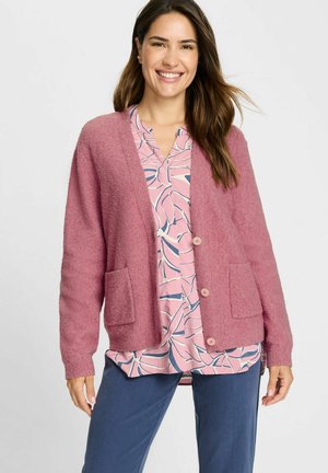 Vezelige roze cardigan met lange mouwen, voorzakken en knoopsluitingen, gelaagd over een bloempatronen blouse en gecombineerd met blauwe broeken.