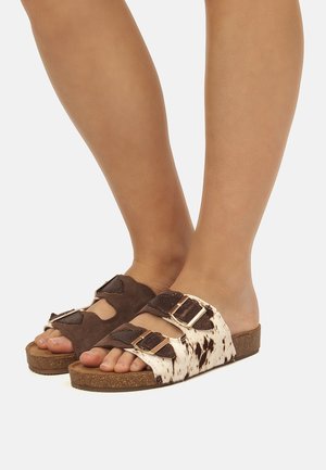 Piedi con sandali a punta aperta con suole in sughero, doppi cinturini e fibbie dorate; un sandalo è marrone scuro, l'altro bianco con macchie marroni.