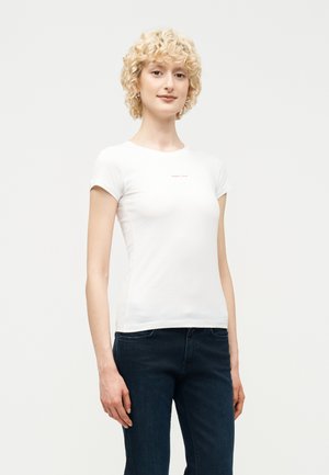 Fiatal nő göndör szőke hajjal, fehér szűk rövid ujjú pólót és sötétkék farmert viselve, sima háttér előtt áll.