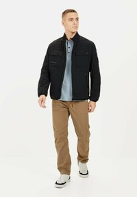 Sort quiltet jakke med ståkrave, frontlommer, parret med en blå polo skjorte og beige bukser, iført hvide sneakers.