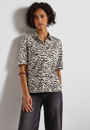 Chemise à manches courtes en tissu beige avec un motif abstrait noir, dotée d'un col et d'une coupe décontractée, associée à un jean sombre.