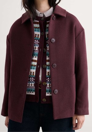 Personne portant une veste en laine bordeaux sur un cardigan en tricot à motifs et une chemise rayée à col, avec un pantalon foncé, sur un fond uni.