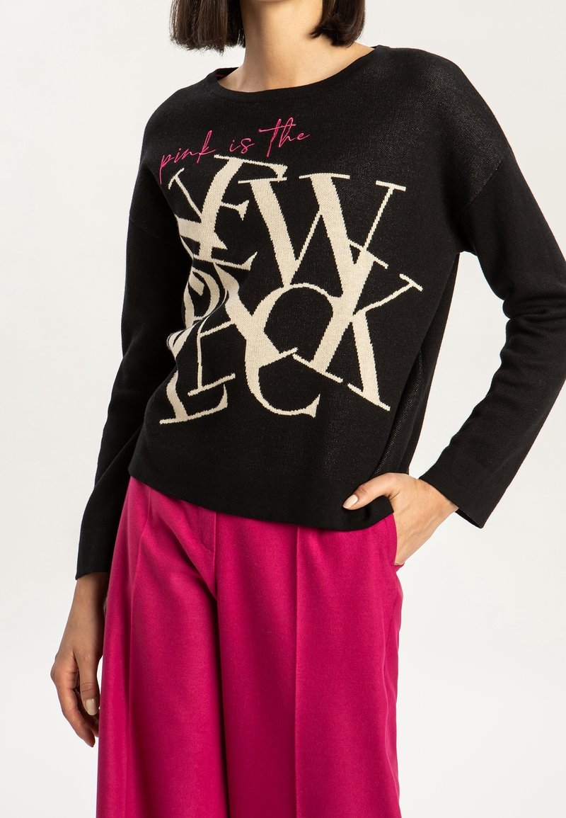 Femme portant un pull noir avec de grandes lettres blanches superposées et un texte rose « pink is the » associé à un pantalon large rose vif.
