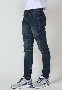 Vaqueros de mezclilla oscuros de corte slim con paneles texturizados y un leve desgaste, combinados con zapatillas deportivas grises con acentos en azul marino.
